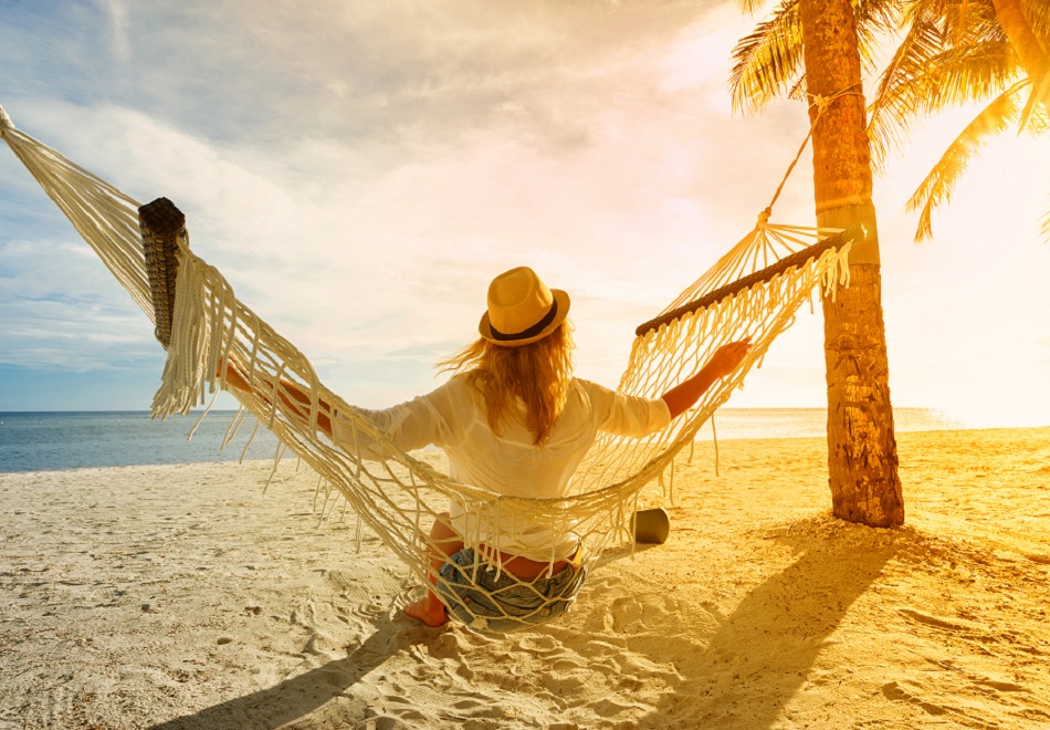 Frau mit Hut sitzt in Hängematte am Strand und schaut auf den Sonnenuntergang