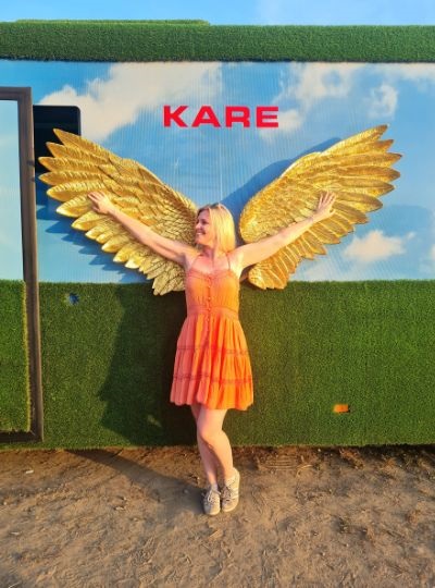 Vor einer Wand mit goldenen Flügeln steht eine Frau im Sommerkleid und breitet die Arme aus